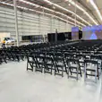 A large warehouse with rows of black padded folding chairs and a large screen.