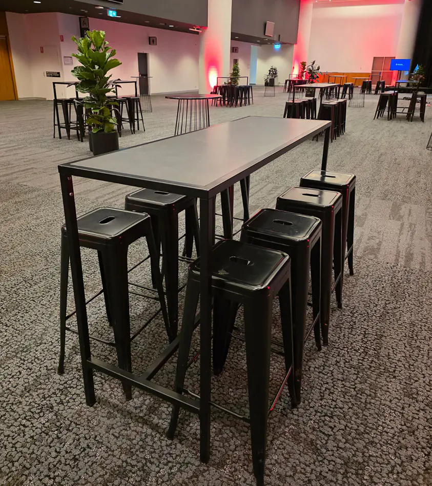 Black rectangular tapas table with industrial black stools positioned inside a large contemporary corporate event venue