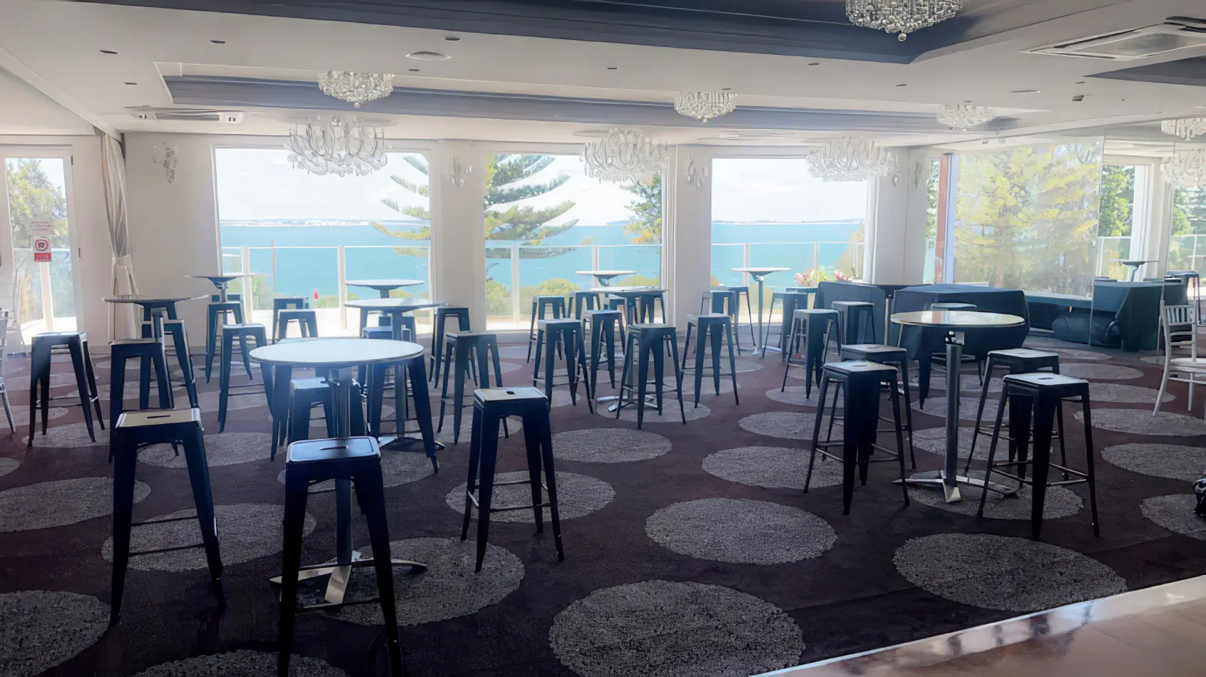 Black tolix stool hire setup inside a bright function room with round cocktail tables, large windows and chandelier lighting overlooking coastal views