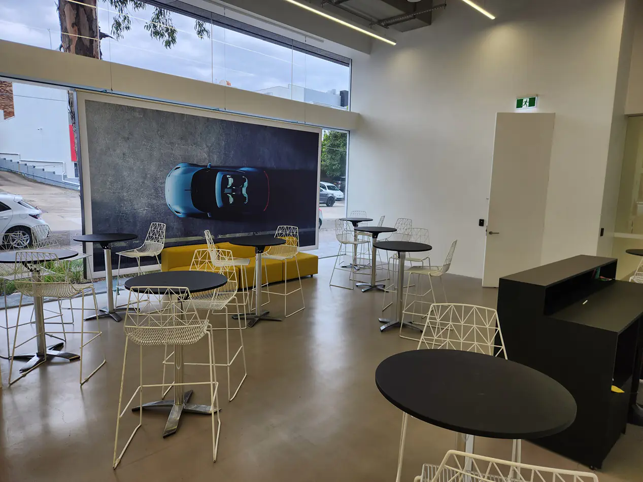 Black top cocktail tables paired with white wire chairs in a modern showroom event space with large windows, polished concrete floor, and contemporary interior design