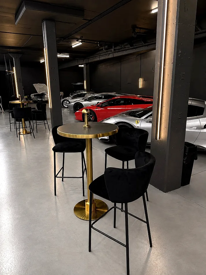 Brass cocktail bar table with a black top surrounded by black velvet bar stools in a supercar showroom.