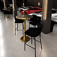 Brass cocktail bar table with a black top surrounded by black velvet bar stools in a supercar showroom.