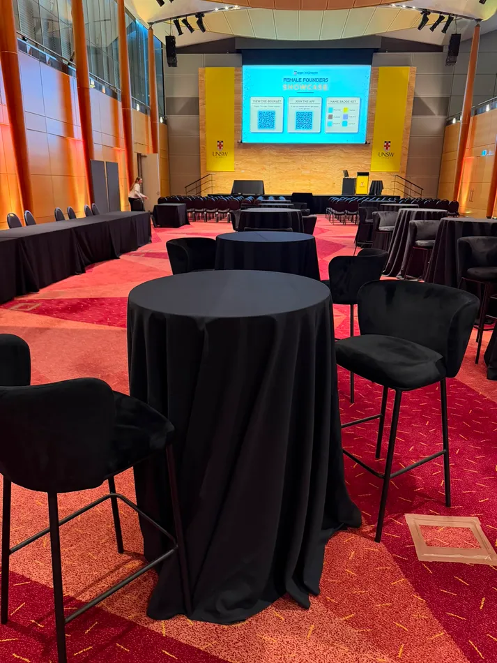 Black velvet bar stools with curved padded backs positioned around black linen cocktail tables at a university conference showcase