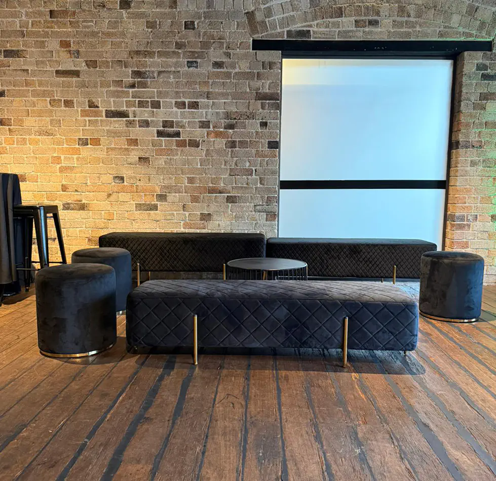 Black velvet bench seating with matching velvet ottomans and round wire coffee table in a warehouse-style event lounge with exposed brick walls