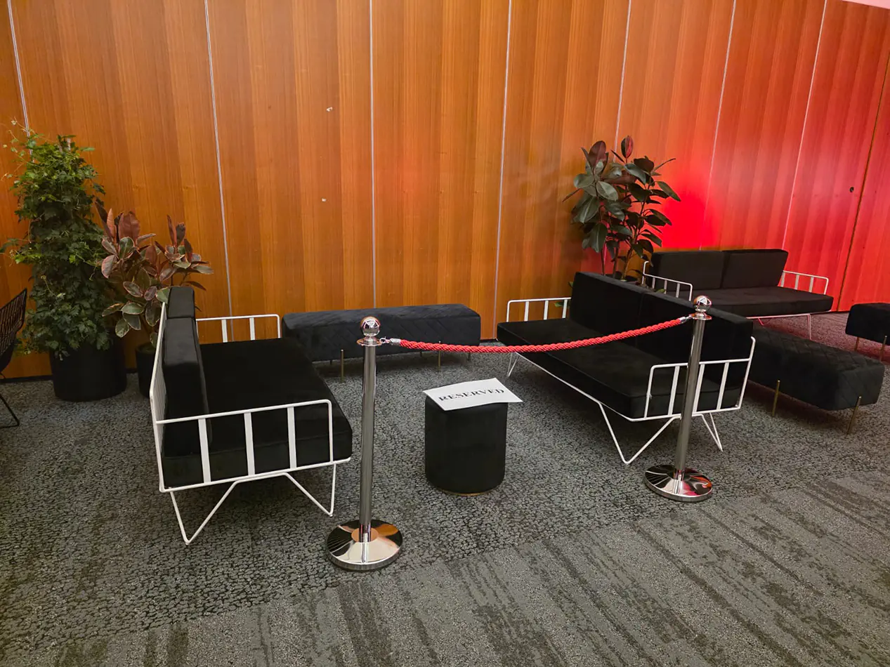 Black velvet lounge seating with white frame sofas, ottoman bench seating, rope barriers and indoor plants in a VIP event area