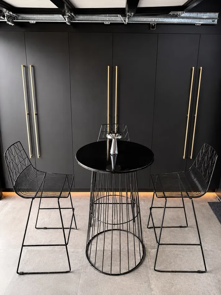 Black wire cocktail table with a gloss black top flanked by two black wire bar stools against a dark panelled wall