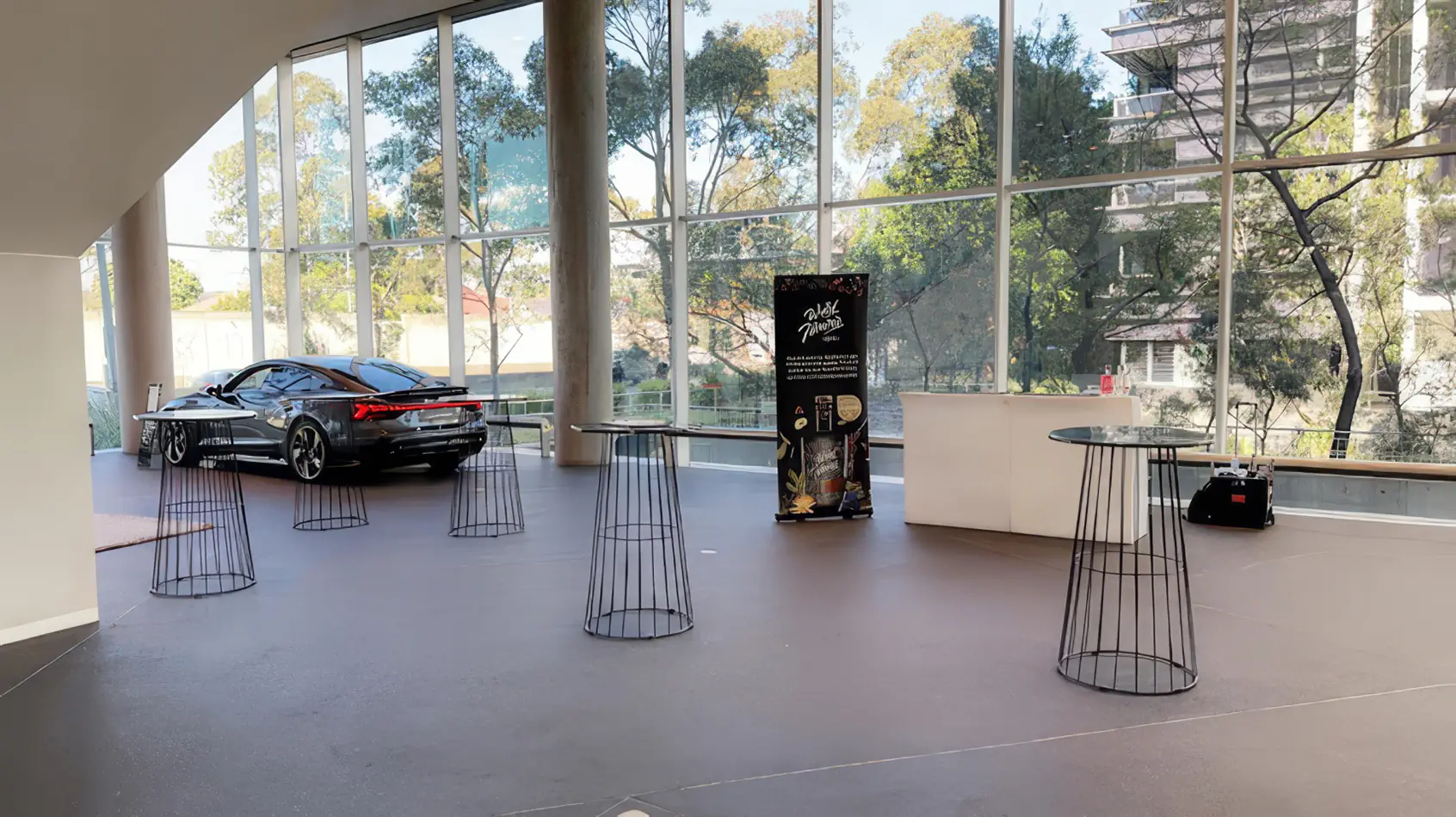Black wire cocktail tables spaced throughout a bright corporate venue with large windows, polished floors and display setup for an event