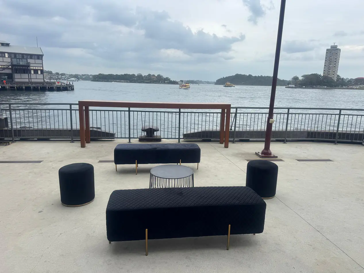 Black wire coffee table with velvet ottoman benches and cylinder stools in an outdoor harbourfront lounge setting