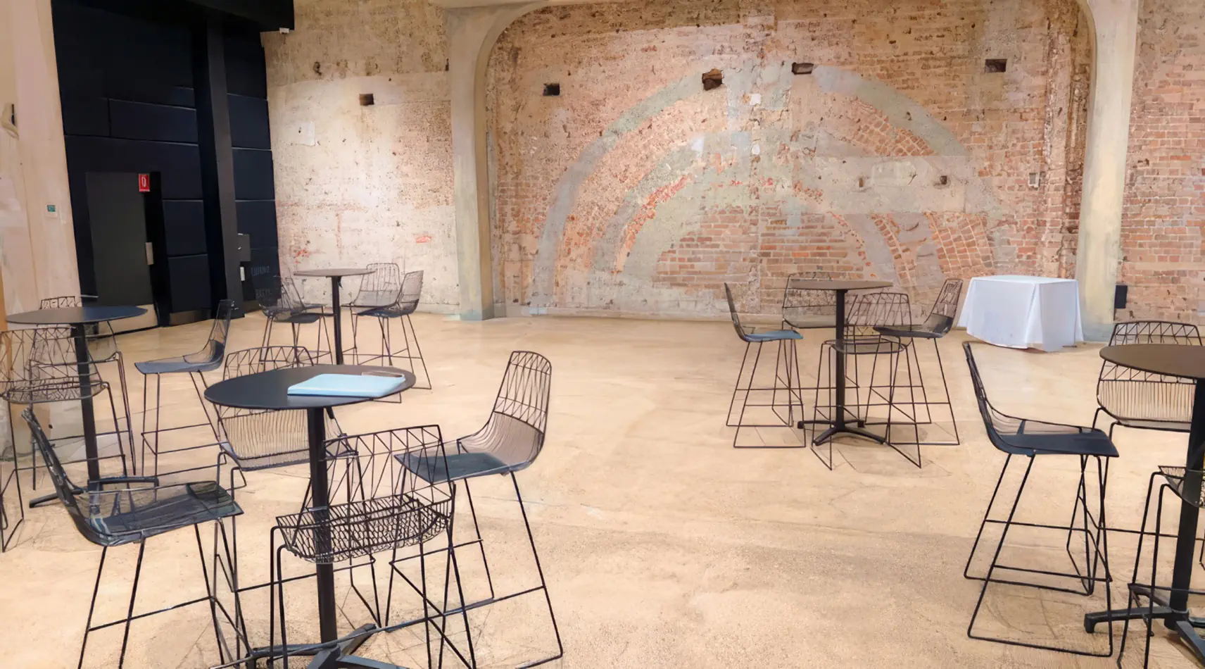 Black wire stools paired with round cocktail tables in an industrial indoor venue with exposed brick walls and polished concrete flooring