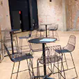 Black wire stools paired with round cocktail tables in an industrial indoor venue with exposed brick walls and polished concrete flooring