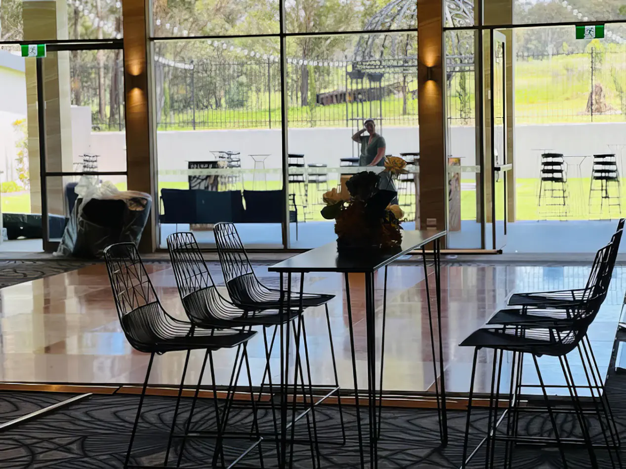 Black wire stools arranged around a high rectangular table inside a modern event space with large glass windows and outdoor garden views