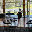 Black wire stools arranged around a high rectangular table inside a modern event space with large glass windows and outdoor garden views