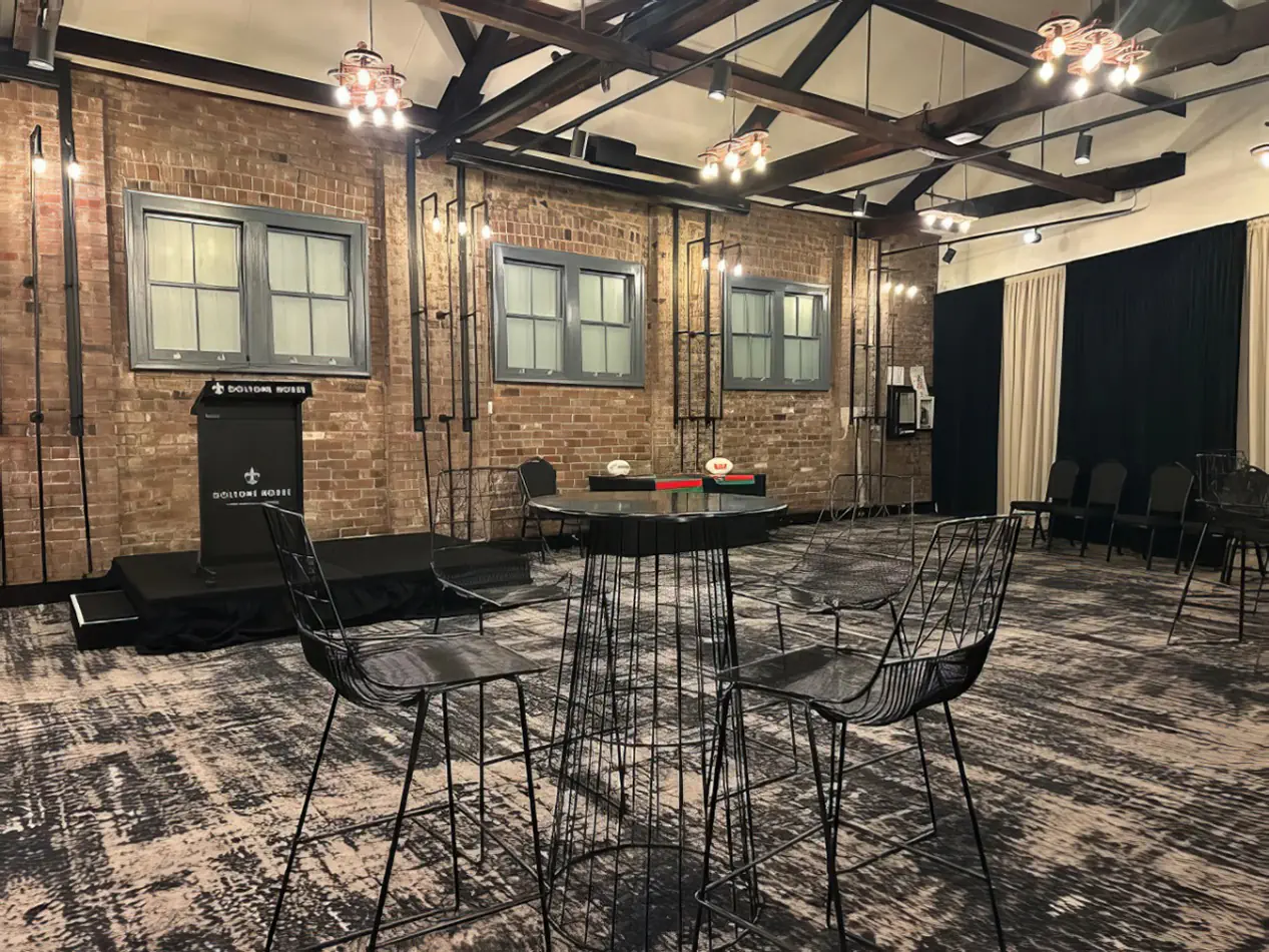 Black wire stools positioned around a round cocktail table in a rustic indoor event room with exposed brick walls and timber ceiling beams