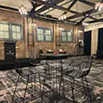 Black wire stools positioned around a round cocktail table in a rustic indoor event room with exposed brick walls and timber ceiling beams