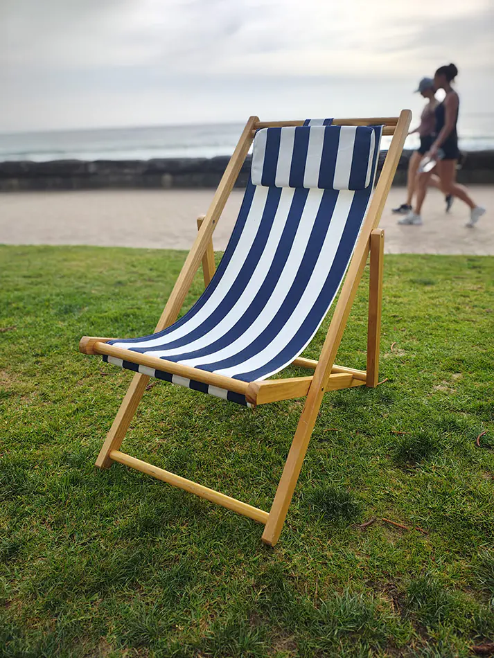 Blue and white striped timber deck chair positioned on grass beside a beachfront promenade and ocean backdrop