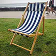 Blue and white striped timber deck chair positioned on grass beside a beachfront promenade and ocean backdrop