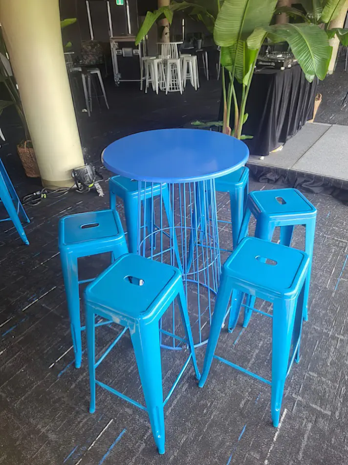 Blue tolix stools arranged around a matching cocktail table in an indoor event space with plants and bar setup in the background
