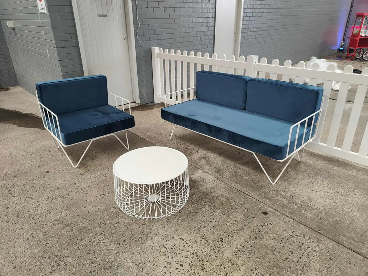 Blue velvet wire lounge chair and sofa set with white wire coffee table arranged in an indoor event space beside white picket fencing