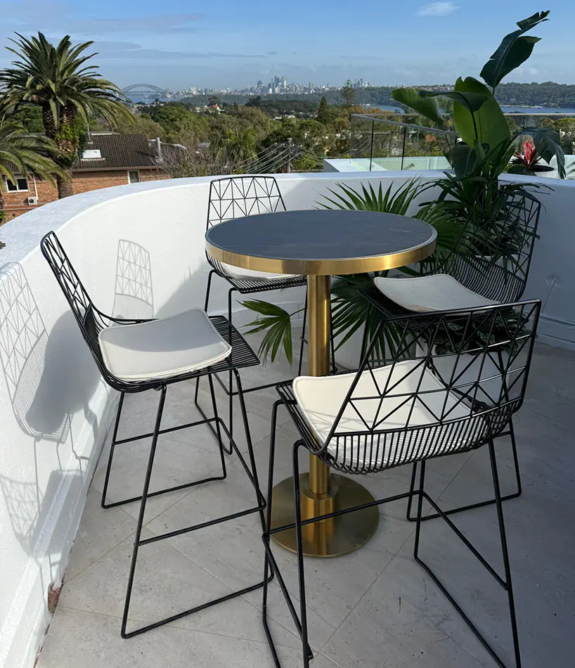 Brass cocktail table with black marble top paired with black wire chairs on a bright balcony overlooking trees and distant city skyline