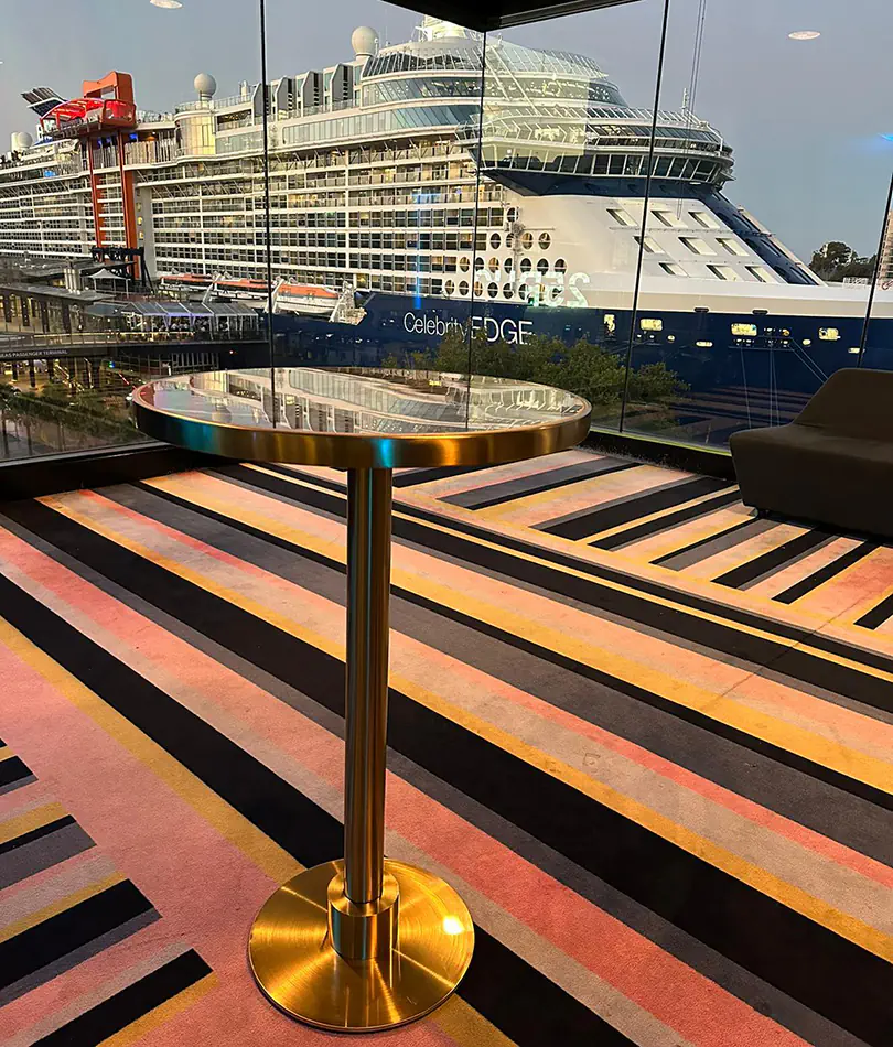 Brass cocktail table with black top positioned in a modern interior space overlooking a harbour with a large cruise ship visible through floor-to-ceiling windows