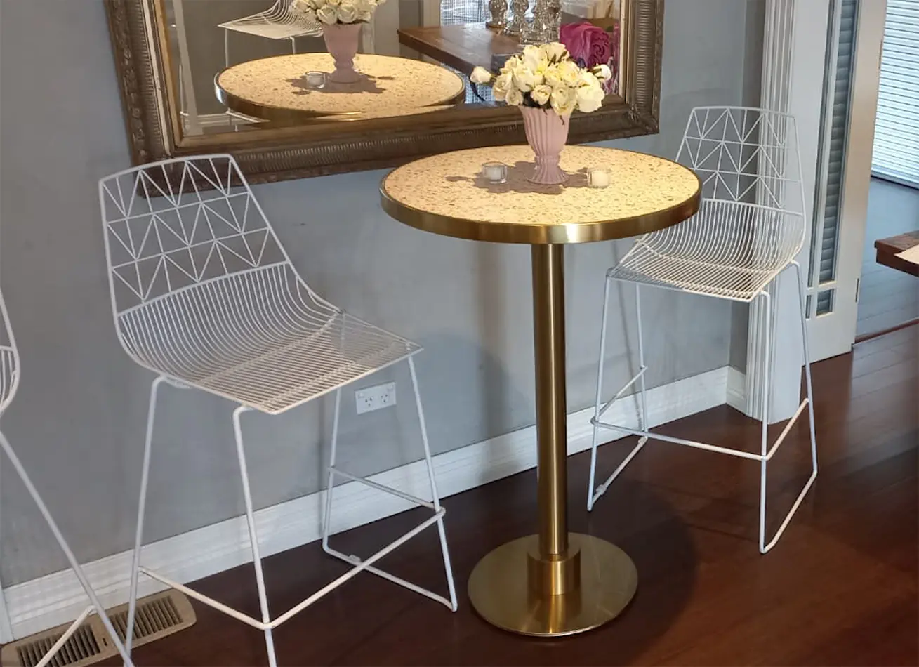 Brass cocktail table with pink terrazzo top styled with white wire chairs, floral arrangement, and decorative mirror backdrop in an indoor setting