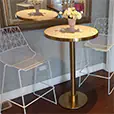 Brass cocktail table with pink terrazzo top styled with white wire chairs, floral arrangement, and decorative mirror backdrop in an indoor setting