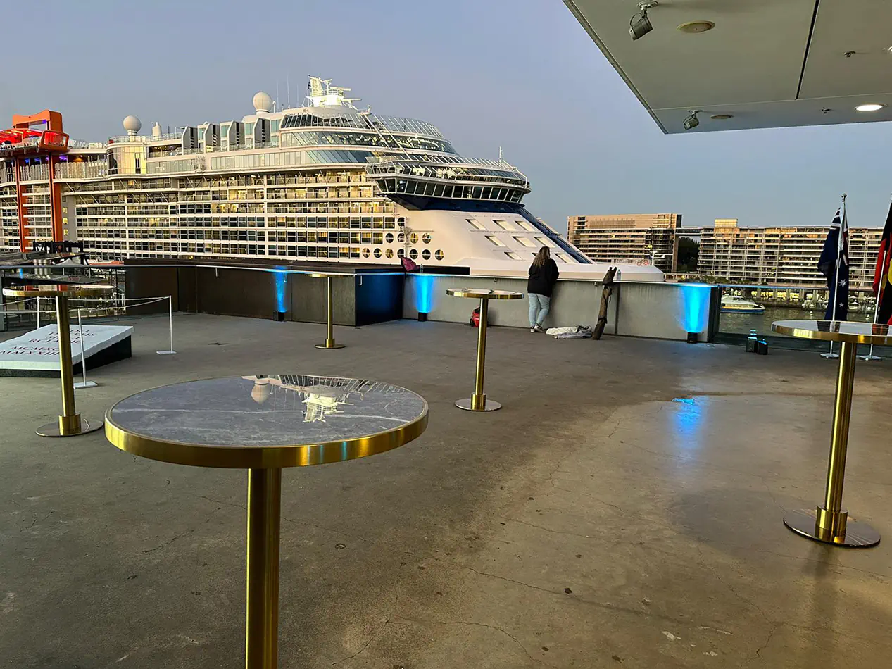 Brass cocktail tables with blue marble tops arranged on a rooftop terrace overlooking a harbour with a large cruise ship and city buildings in the background