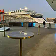Brass cocktail tables with blue marble tops arranged on a rooftop terrace overlooking a harbour with a large cruise ship and city buildings in the background
