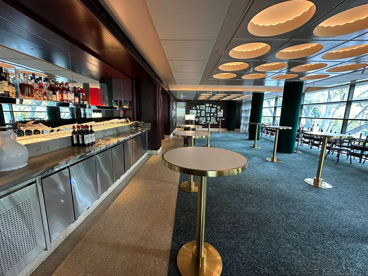 Brass cocktail tables with white tops arranged throughout a modern bar venue interior with marble bar counter, shelving, and ambient ceiling lighting