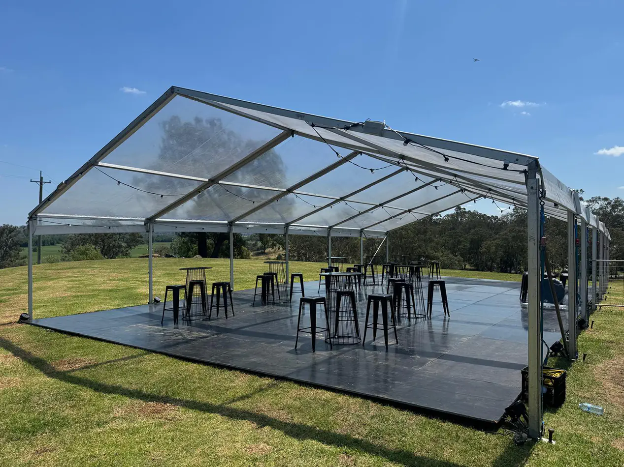 Clear roof marquee set up on grass with black flooring, festoon lighting and bar tables arranged for an outdoor event in a rural setting