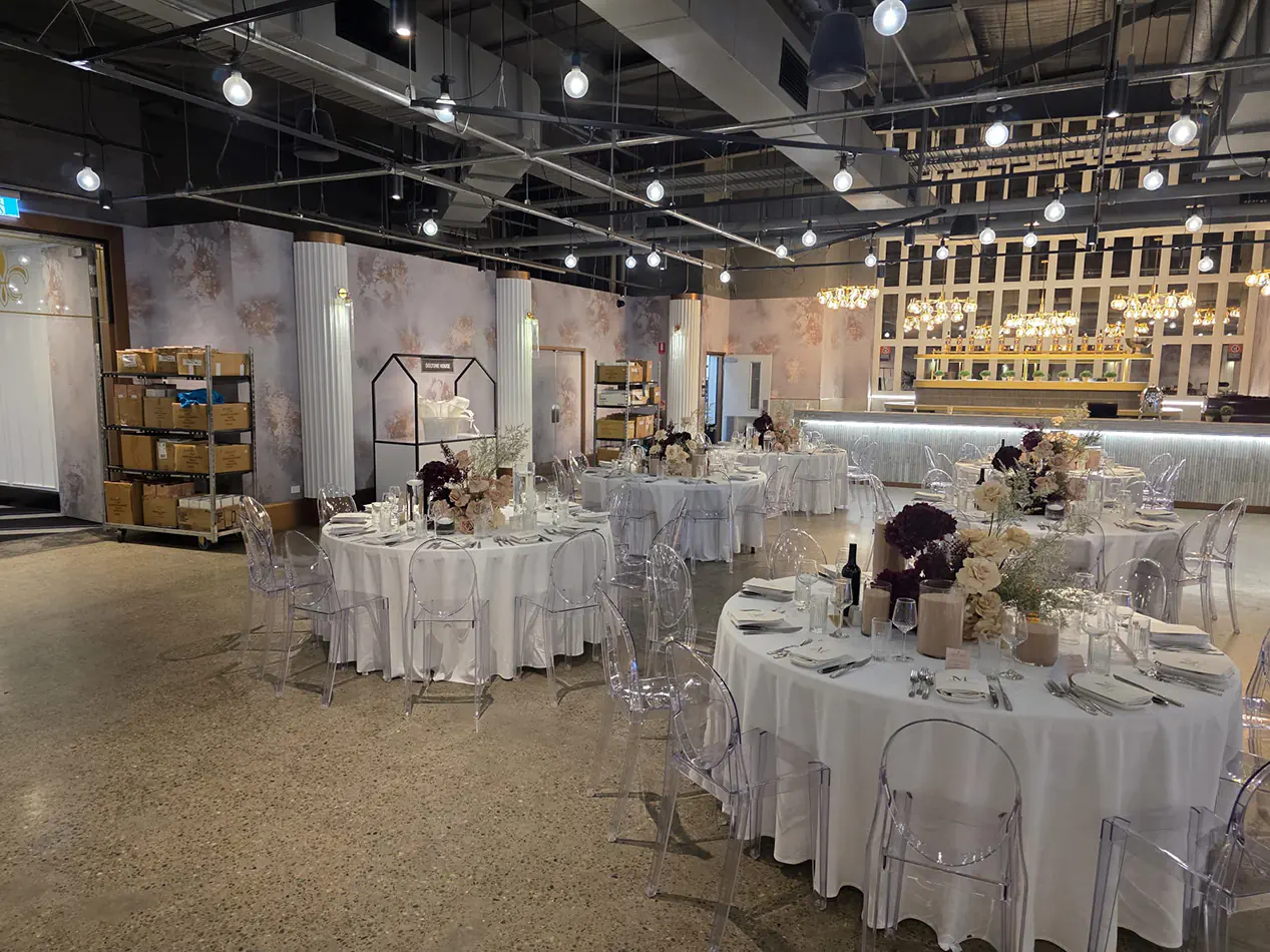 Modern wedding venue with round banquet tables, clear Victorian chairs, floral arrangements and decorative lighting throughout the reception space
