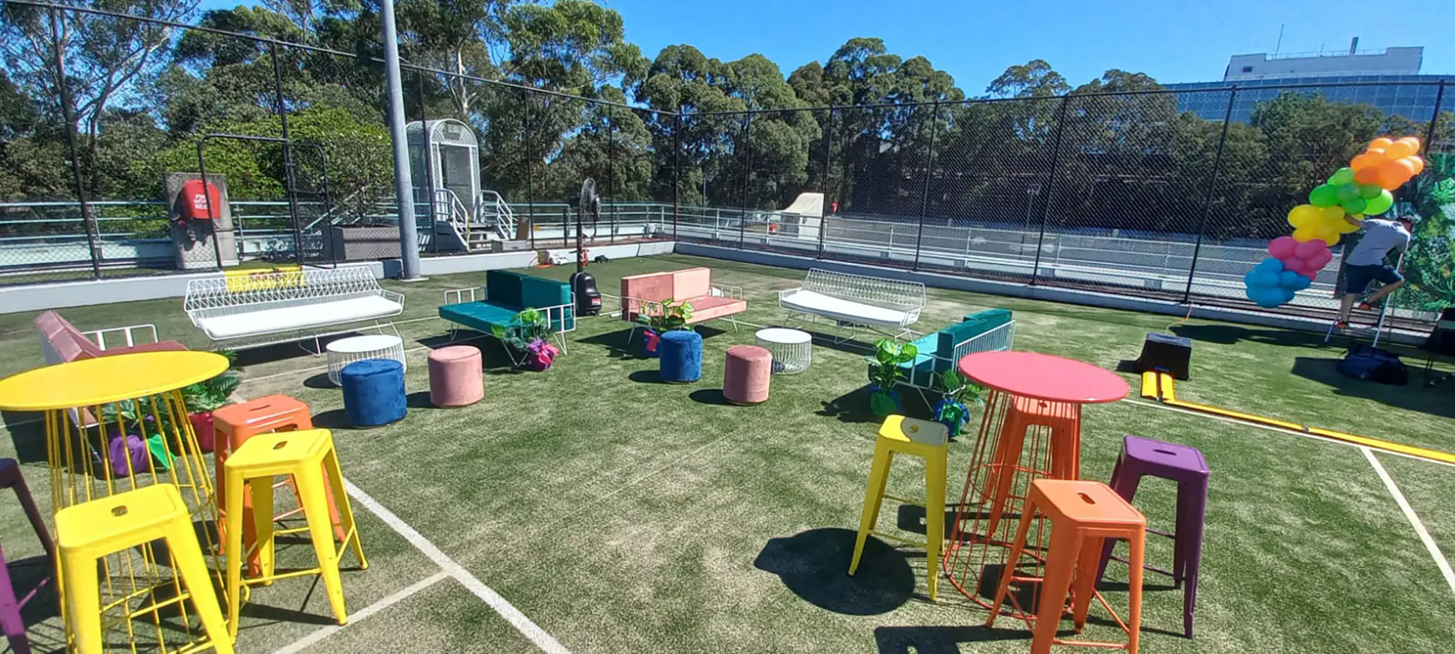 Colourful outdoor event setup with wire cocktail tables, bar stools, lounge seating and decorative elements on artificial grass