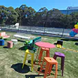 Colourful outdoor event setup with wire cocktail tables, bar stools, lounge seating and decorative elements on artificial grass
