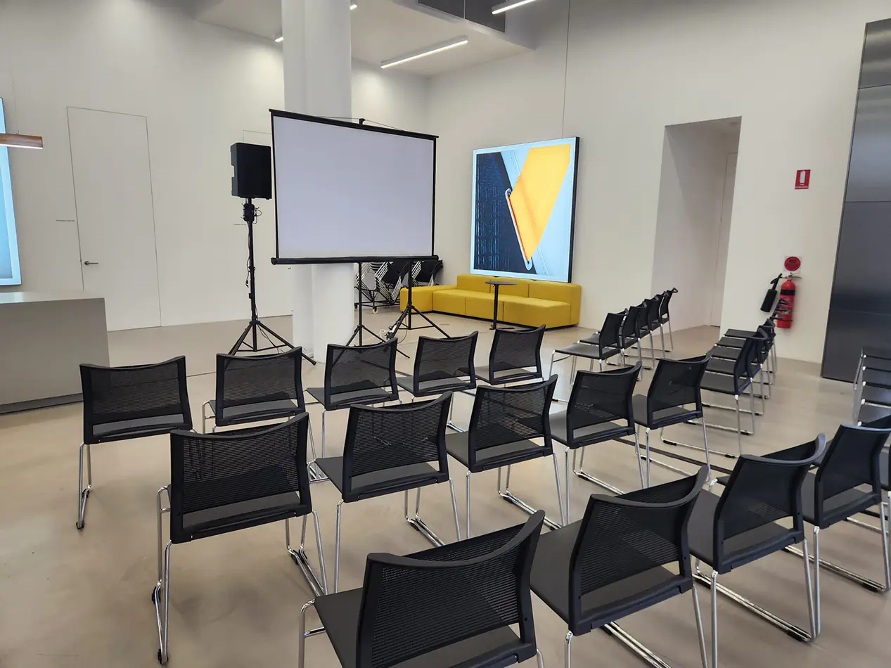 Corporate conference setup with projector screen, PA speaker system, and rows of black presentation chairs inside a modern venue