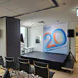 Conference stage with raised platform, speakers on stands, branded backdrop, and seating arranged for corporate presentation in office space