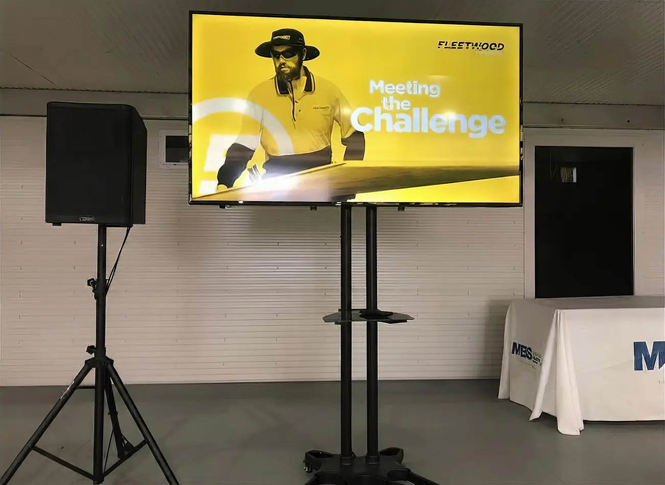 Corporate TV hire setup with a large screen on a stand, PA speaker, presentation table and branded event slide displayed on screen
