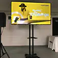 Corporate TV hire setup with a large screen on a stand, PA speaker, presentation table and branded event slide displayed on screen