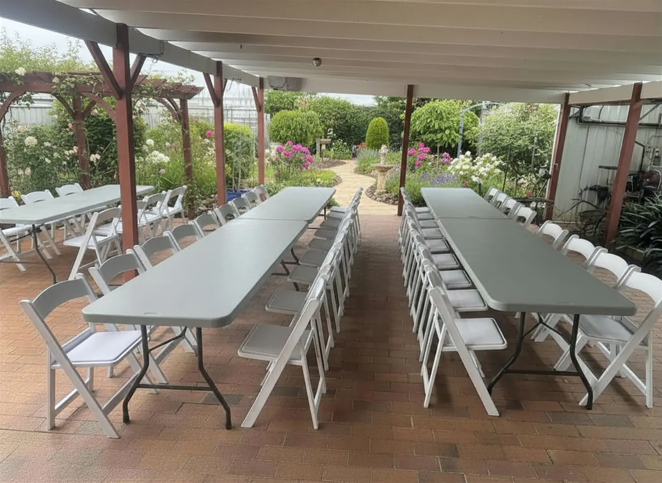 Covered patio event setup with grey plastic trestle tables and white folding chairs arranged for a garden gathering