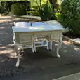 Cream vintage table with decorative handles and curved legs, positioned with two white chairs on a paved garden path surrounded by greenery.