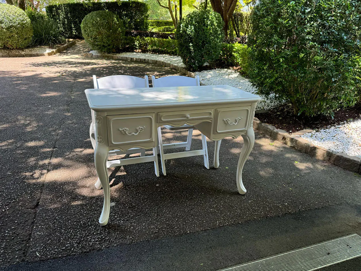 Cream vintage table with decorative handles and curved legs, positioned with two white chairs on a paved garden path surrounded by greenery.