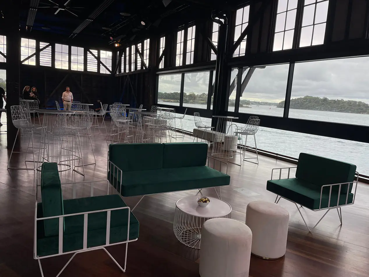 Emerald green velvet lounge seating with white wire frame furniture and ottoman stools inside a waterfront event venue with floor-to-ceiling windows