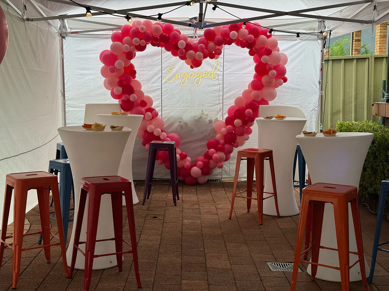 Cocktail tables with white covers and colourful bar stools set up under a marquee with pink balloon arch and engagement sign backdrop