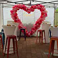 Cocktail tables with white covers and colourful bar stools set up under a marquee with pink balloon arch and engagement sign backdrop