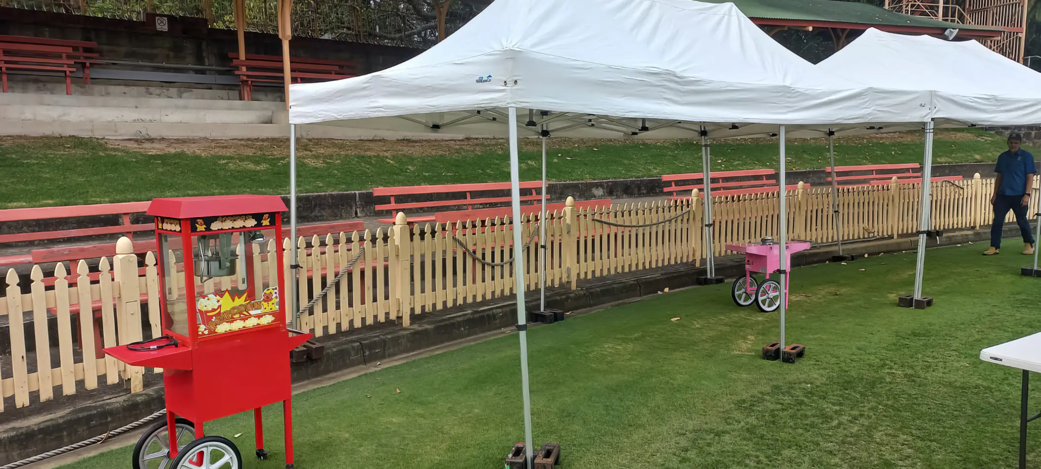 Popcorn machine cart in bright red set up under outdoor canopy tents alongside fencing and event equipment