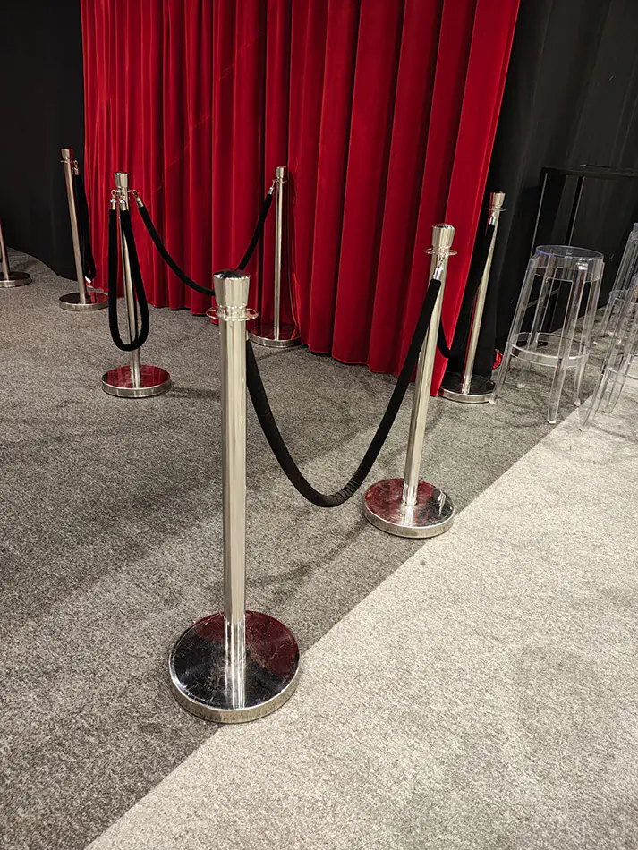 Event queue barriers with chrome stanchions and black ropes set in front of a red curtain backdrop on carpeted floor
