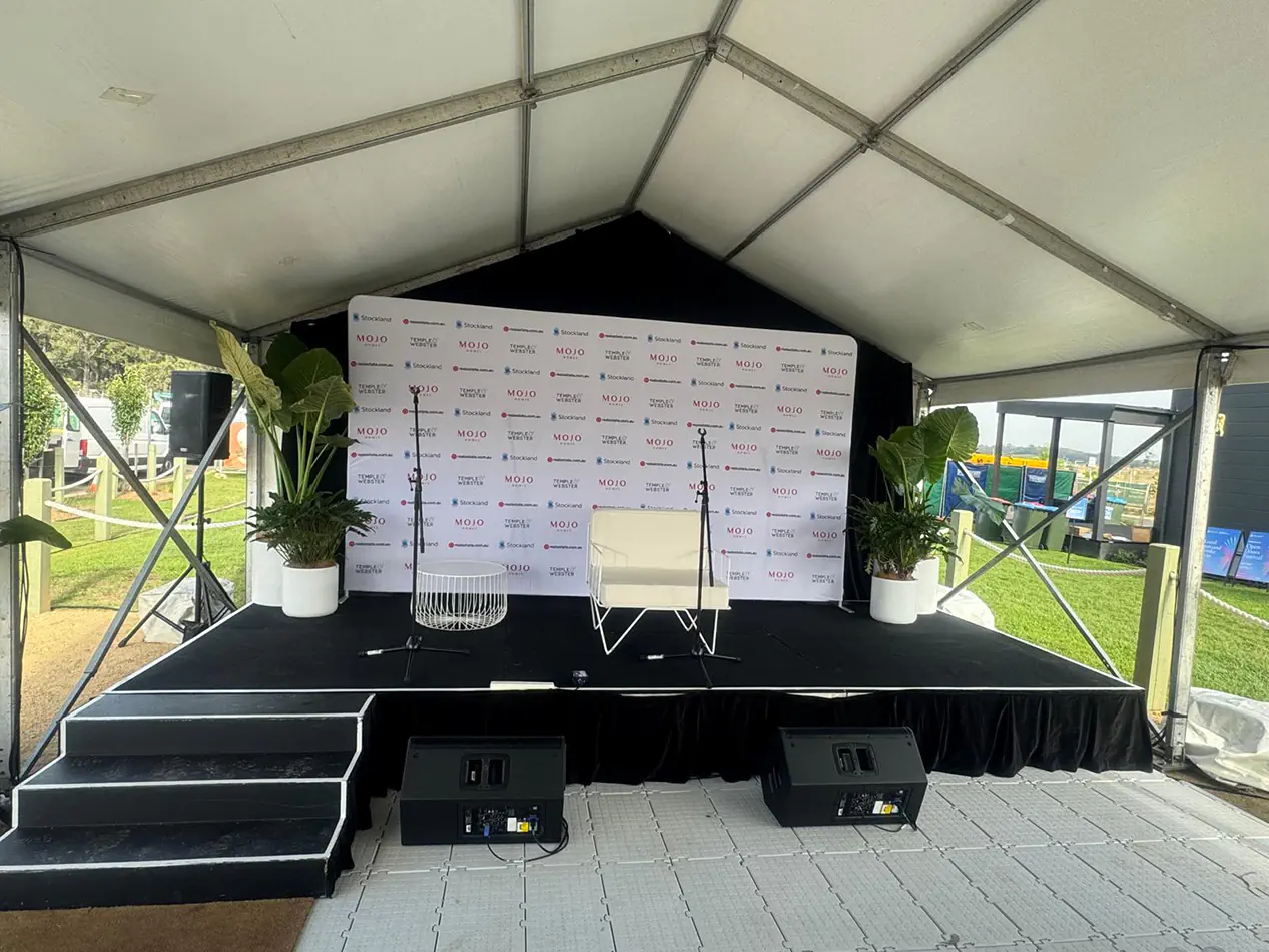 Event stage setup with PA speakers, microphones, branded media backdrop, and presentation area for a live corporate event