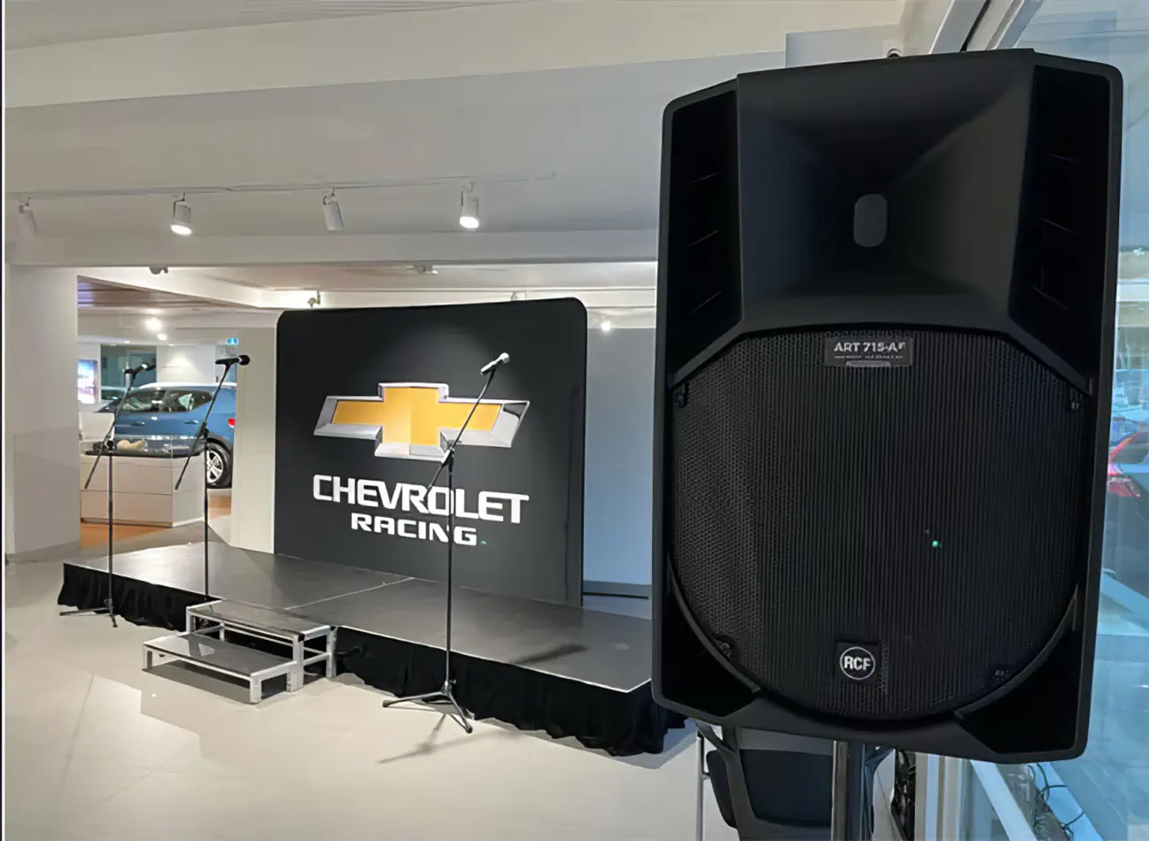 Event stage setup with black platform, microphones on stands, large PA speaker in foreground, and Chevrolet Racing display backdrop in showroom environment