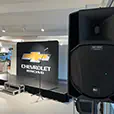 Event stage setup with black platform, microphones on stands, large PA speaker in foreground, and Chevrolet Racing display backdrop in showroom environment