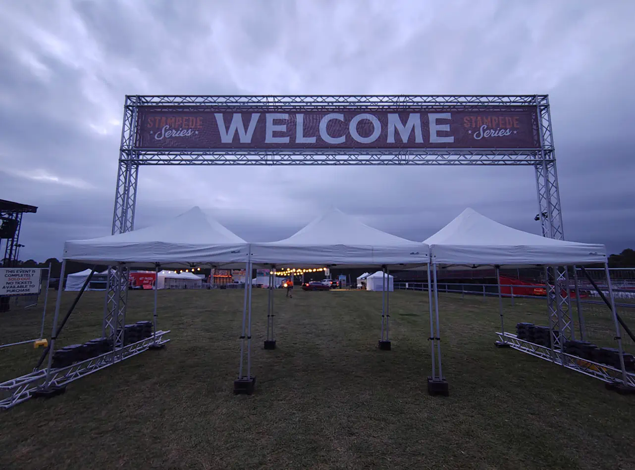 White pop-up marquees positioned beneath a large festival welcome truss entrance at an outdoor event venue