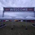 White pop-up marquees positioned beneath a large festival welcome truss entrance at an outdoor event venue